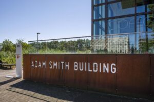 Vooraanzicht van het Adam Smith Building aan de Thomas R. Malthusstraat met moderne glazen gevel en roestbruine wand met witte belettering.